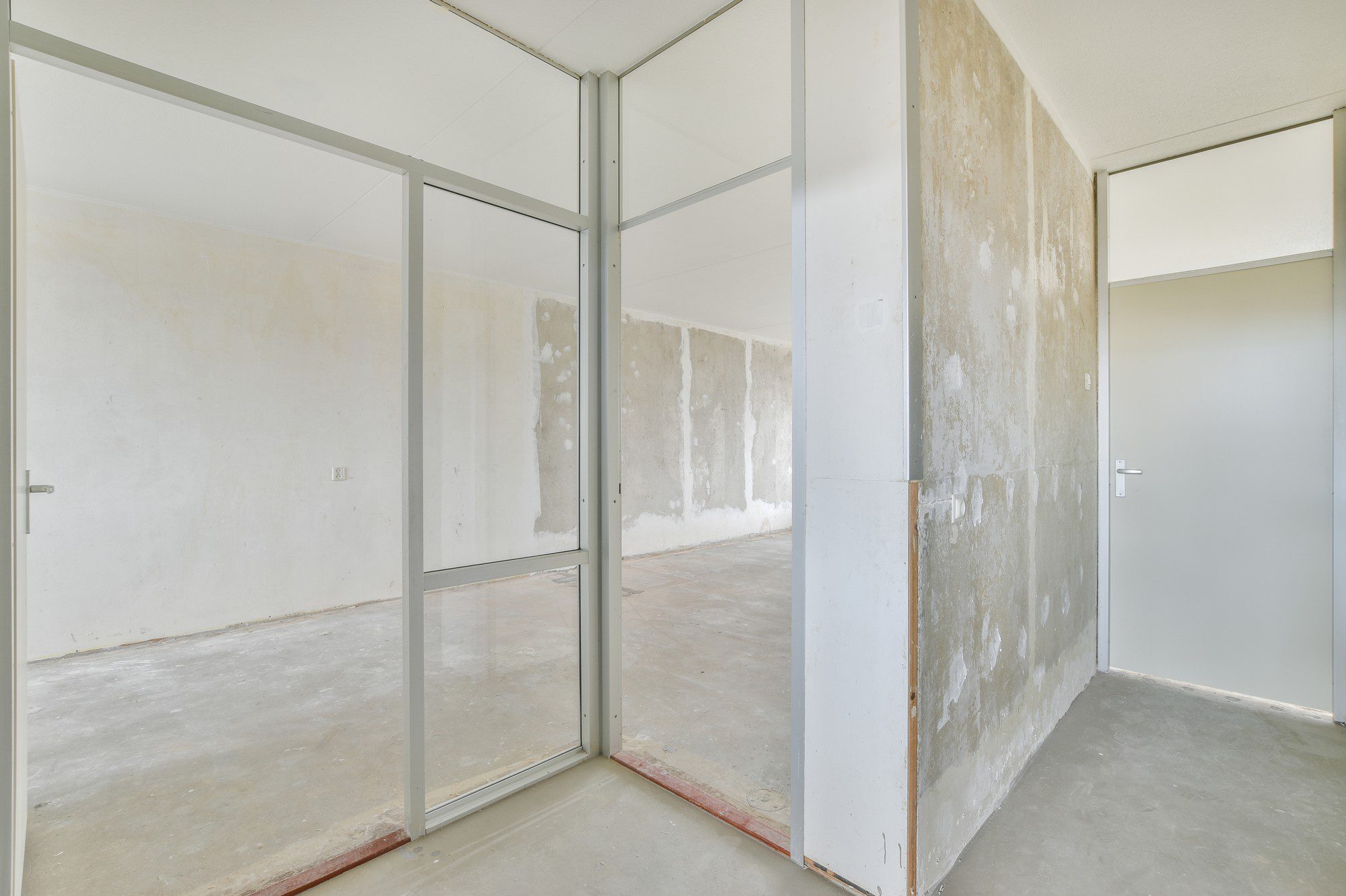 The image depicts an interior space that is in an unfinished or renovation state. There is a glass partition with a metal frame, dividing the space into separate areas. The walls look like they've been stripped of wallpaper or old paint, with some areas showing signs of spackling or patching, which is typically done before repainting or application of new wall coverings. The floor is bare concrete, and on the right, there's a closed door with a handle, suggesting an entry into another room or closet. The room is empty, suggesting that it is likely under construction, renovation, or refurbishment. The overall appearance is that the space is in the midst of a renovation project, awaiting further finishing work.