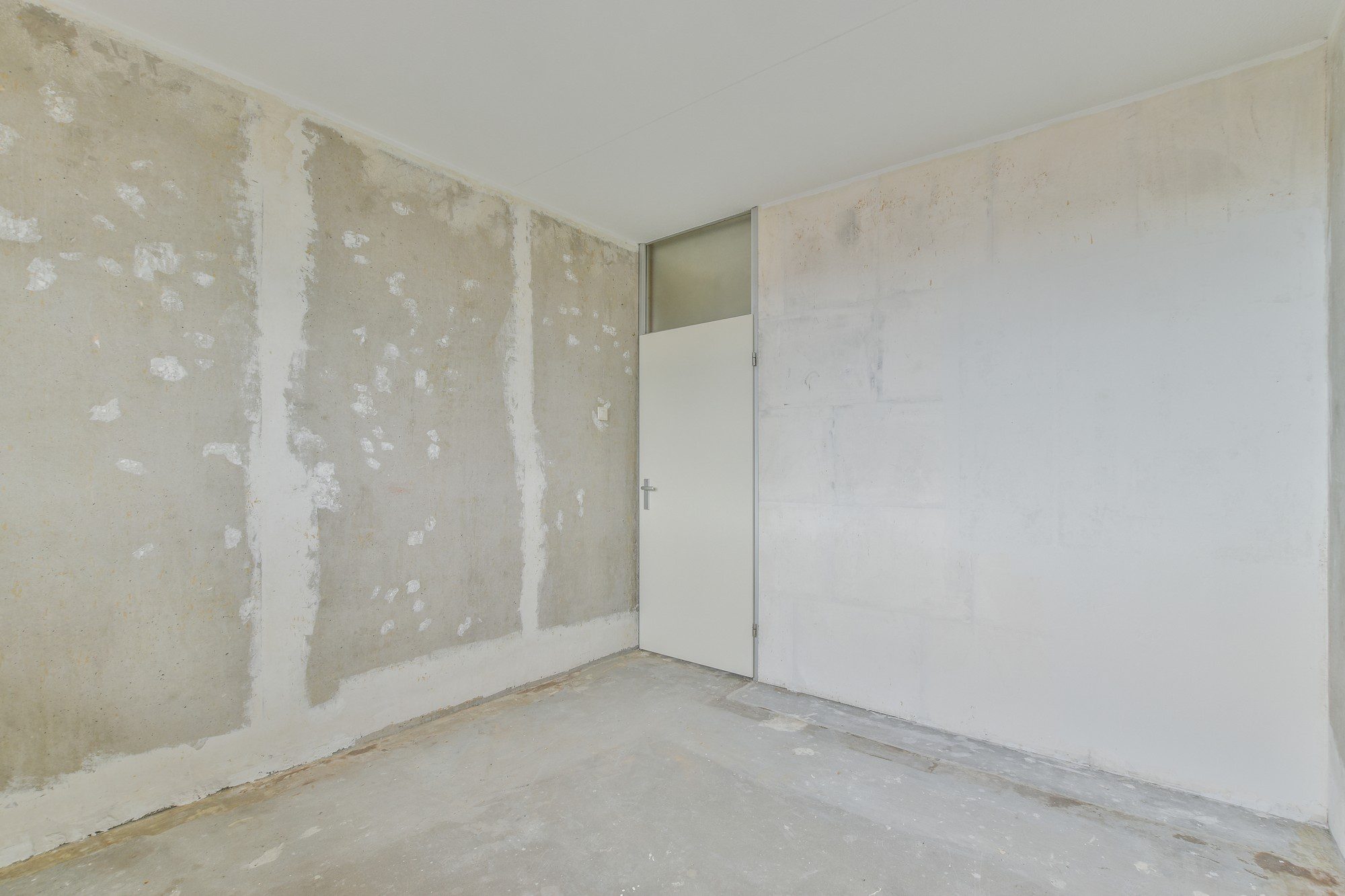 This is an image of an empty room. The walls look to be under construction or renovation, as they are partially stripped or unfinished, exposing the concrete beneath. The wall on the left and the back appears to have had some kind of plaster or wall finish which is now flaking off or has been scraped off in certain areas. There's a closed door on the wall that appears to be in relatively good condition compared to the walls. The ceiling seems finished and painted white, but the floor is bare concrete with some debris or residue on it. The room gives an overall appearance of being in a state of disrepair or in the midst of refurbishment.