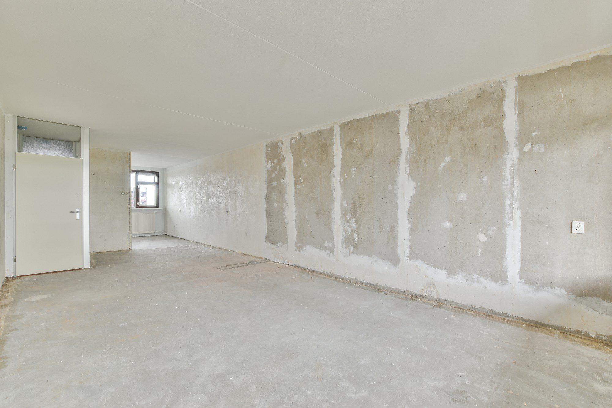 The image shows an empty room that appears to be under renovation or preparation for renovation. The floor is bare concrete without any carpeting or other flooring material installed. The walls are stripped to what looks like bare concrete or plaster as well, with patches of filler or spackle indicating that holes or imperfections have been filled in preparation for painting or wallpapering.

There's a window on the far side providing natural light. On the left, there is a door that is open, leading to another room or space; above this door, a small transom window can be seen. The room has a flat ceiling with no visible light fixtures, and there are some electrical outlets on the walls. The general condition suggests that this space may be part of a construction or remodeling project before it is finished and made livable.