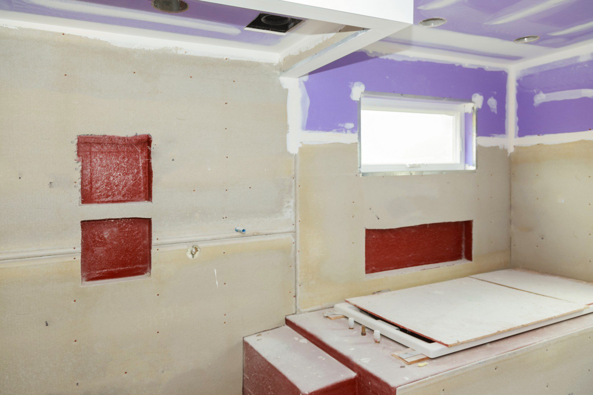 This image shows a room under renovation. The walls are partially stripped, revealing patches of underlying red material that might be a previous layer of wall material or potentially adhesive residue. Portions of drywall are not yet finished, as indicated by visible screw heads and unsealed joints. Purple-coloured spackling or joint compound can be seen applied along the seams and screw holes in the drywall to create a smooth surface upon sanding.

The ceiling has recessed lighting fixtures installed, and there's a small window with a white frame on one wall. The window allows daylight to filter into the room. To the right, there is a built platform or step, possibly indicating a design feature or future installation area, which seems to be covered with protective dust sheets. The room appears dusty and in a transitional state, typical of construction sites or renovation projects before the final finishing touches are applied.