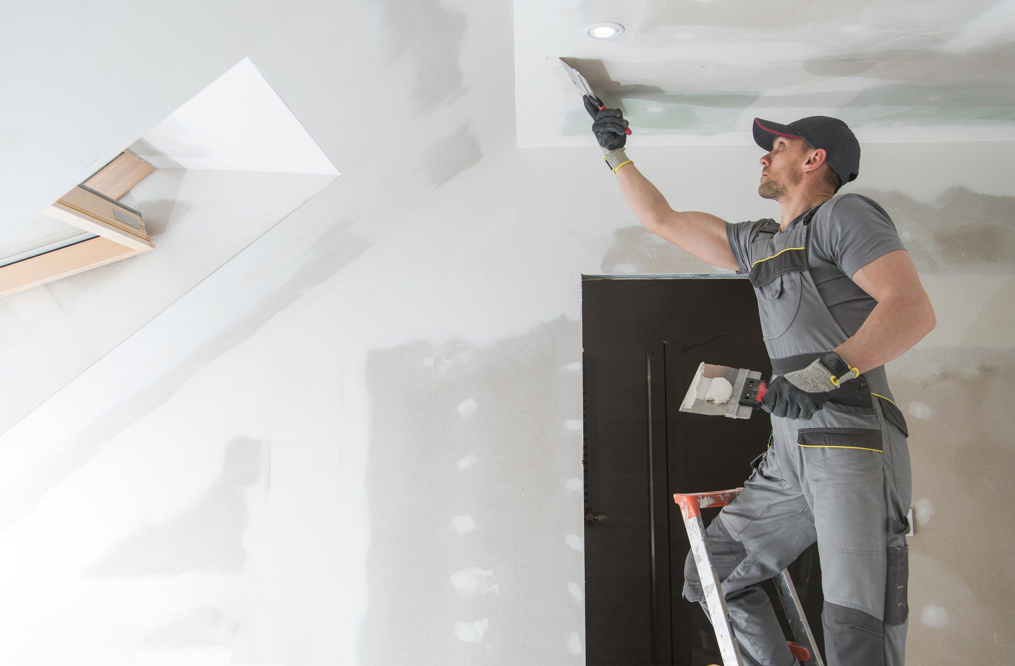 The image shows a person working on a drywall installation or repair. The person is likely a professional construction worker or handyman. They are standing on a stepladder and using a spatula or putty knife to apply joint compound (often referred to as mud) to the seams of the drywall. The worker is wearing a grey work uniform with yellow detailing, gloves, a cap, and a tool belt. There is also a portable light on the ceiling and a skylight in the room. The wall behind the worker is partially finished, with some of the drywall taped and mudded and others just installed and not yet smoothed over.