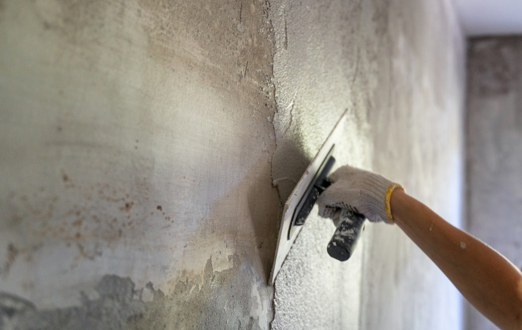 This image shows a person's hand, wearing a white knitted glove with a yellow wristband, applying plaster to a wall with a plastering trowel. The process is likely part of wall construction or repair, and the person is smoothing out the plaster to create a flat surface for finishing. The wall appears to be in a building under renovation or construction.