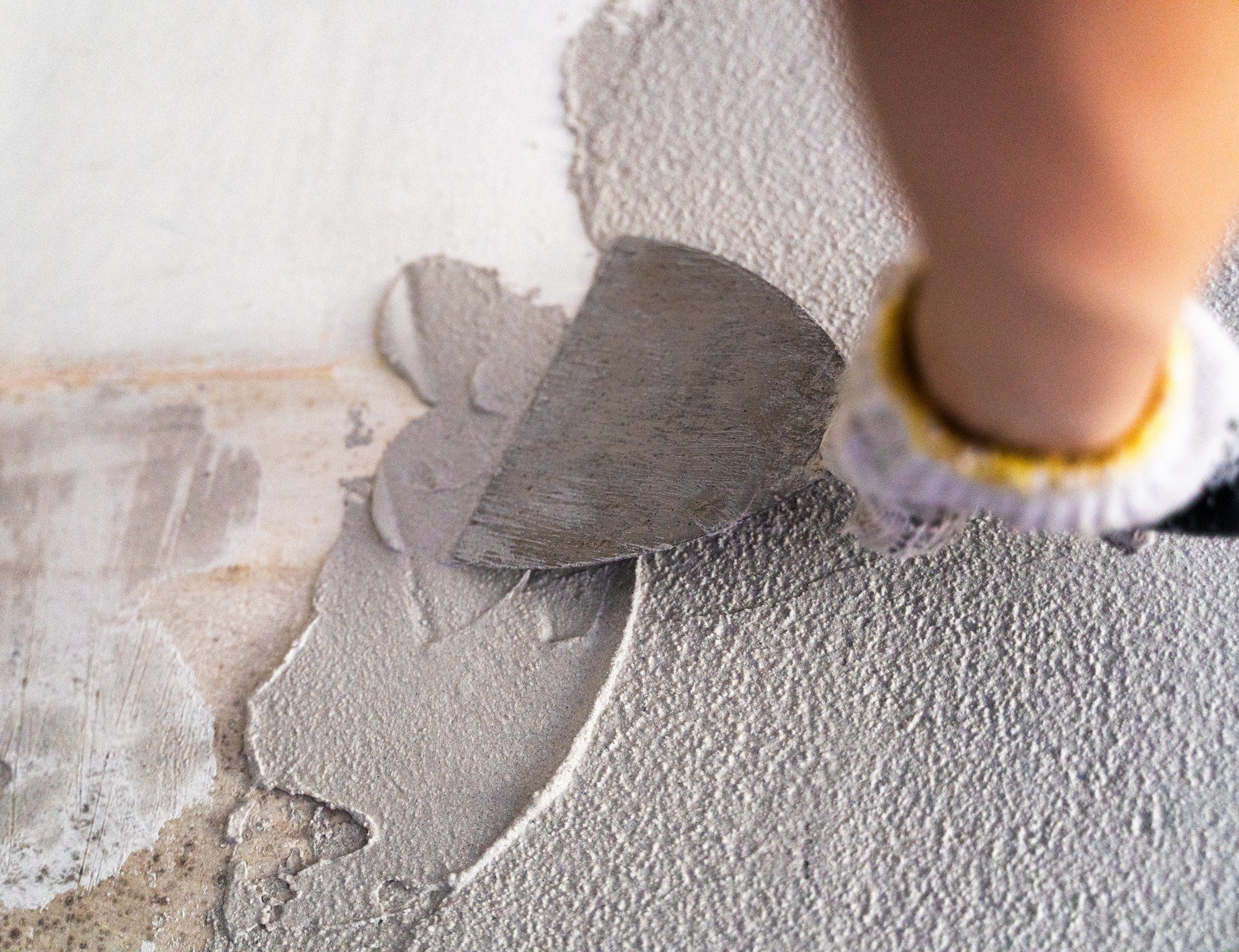 This image depicts a person applying a grey compound to a wall using a trowel. The compound is likely plaster or joint compound, and it is being spread to either repair or smooth the surface of the wall. The person's hand is shown holding the trowel, and they seem to be in the process of skimming the wall. The background indicates that this is part of some larger construction or repair work inside a building.