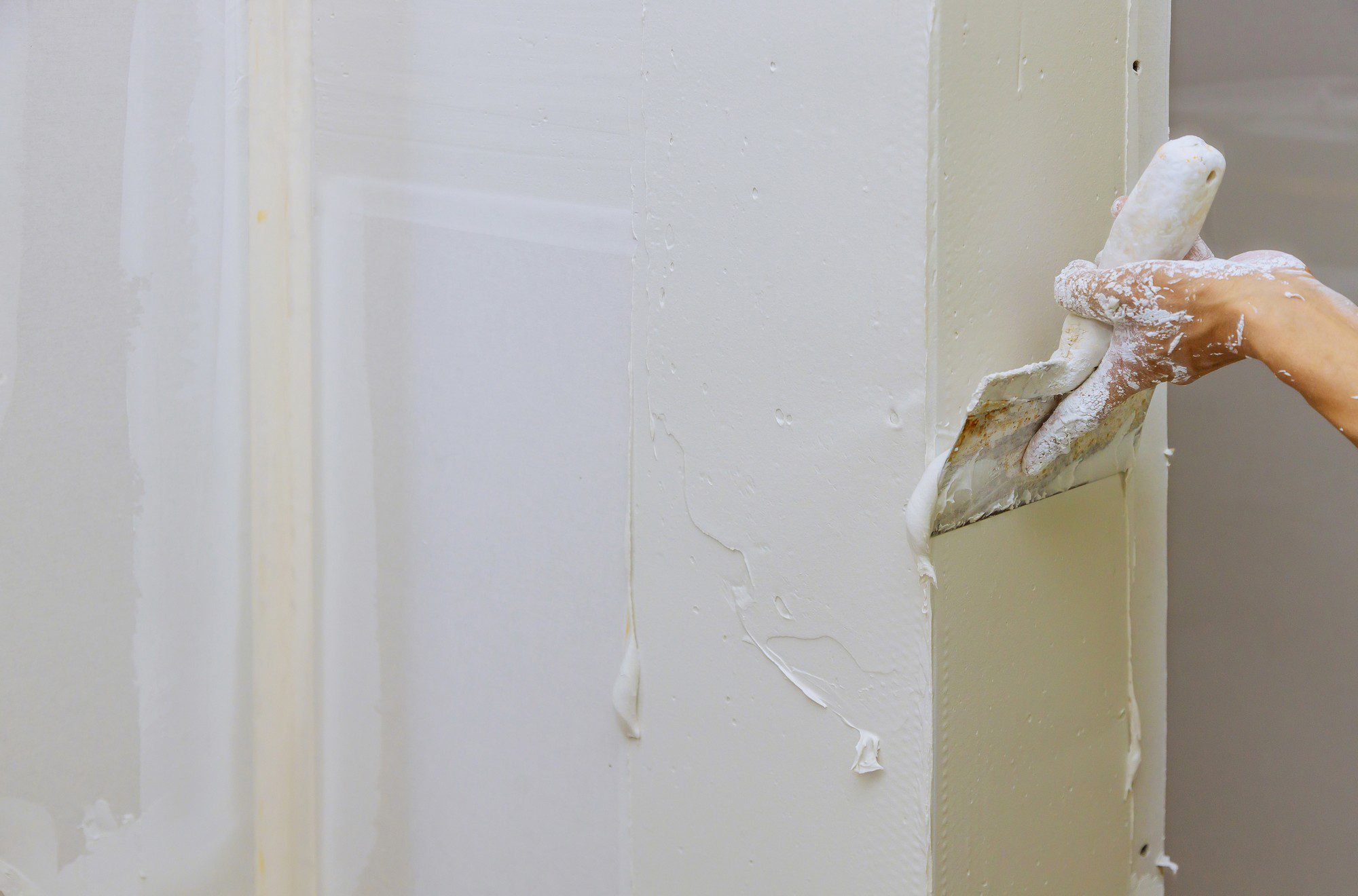 The image shows a person's arm holding a putty knife or scraper, removing what appears to be old, peeling paint from a wall or door frame. The person is wearing a glove covered in white dust, which suggests they are in the process of scraping or sanding a painted surface, likely as part of a refurbishment or redecoration project. The wall has multiple layers of paint, as evidenced by the peeling sections. There are paint chips and dust on the surface as well as on the glove, indicating the physical nature of the work being performed.