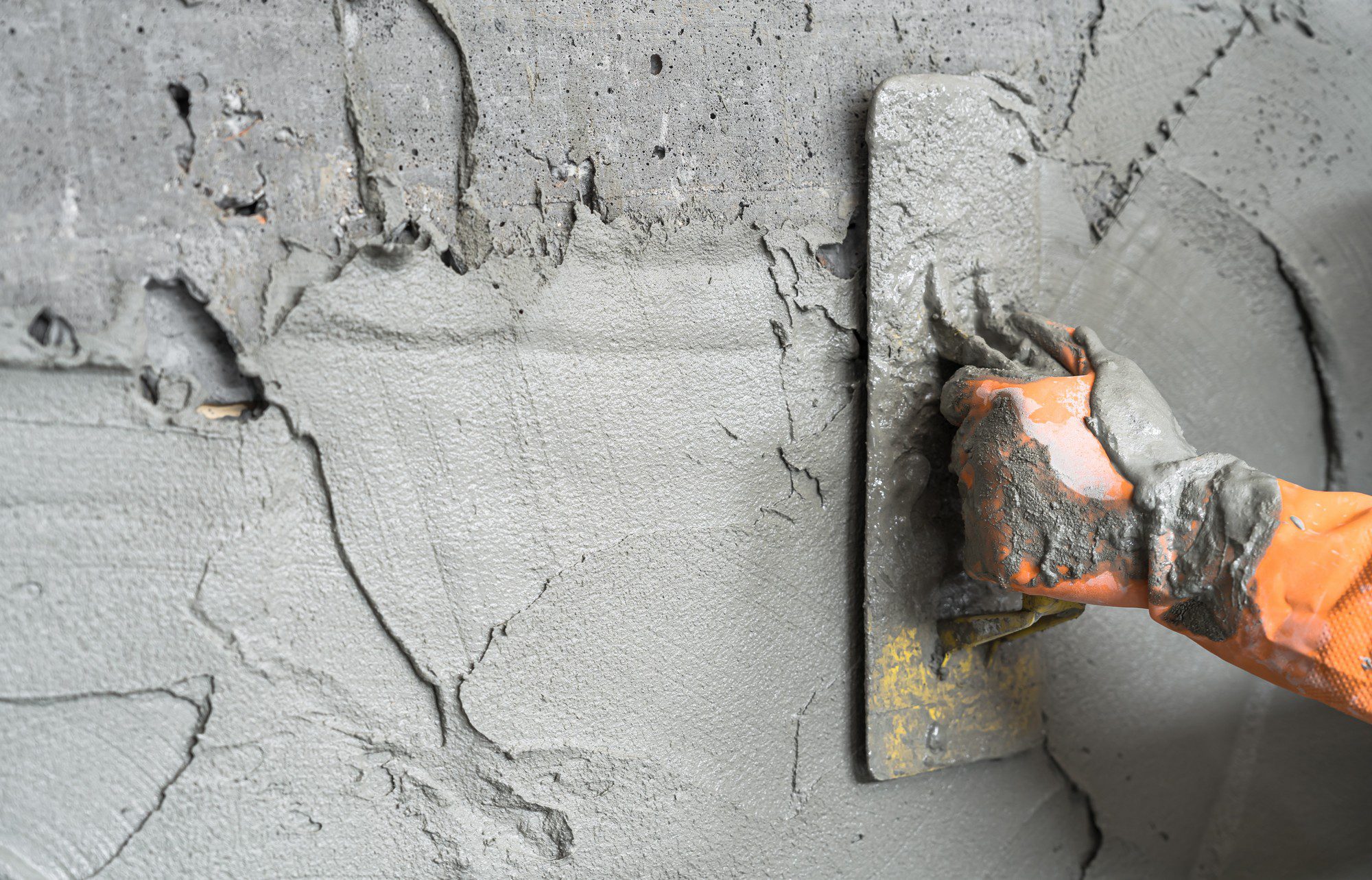 This image shows a close-up of a hand wearing an orange and gray glove, smoothing wet cement or concrete with a trowel. It appears that the person is in the process of applying or finishing the cement on a surface. The texture of the freshly applied cement is rough, and there are visible lines and irregularities indicating that the work is ongoing. The trowel that the person is using has some yellow markings on it, suggesting it may have been used previously for similar tasks. The focus of the picture is on the action of spreading or smoothing the cement, and it provides a glimpse into the process of masonry or construction work.