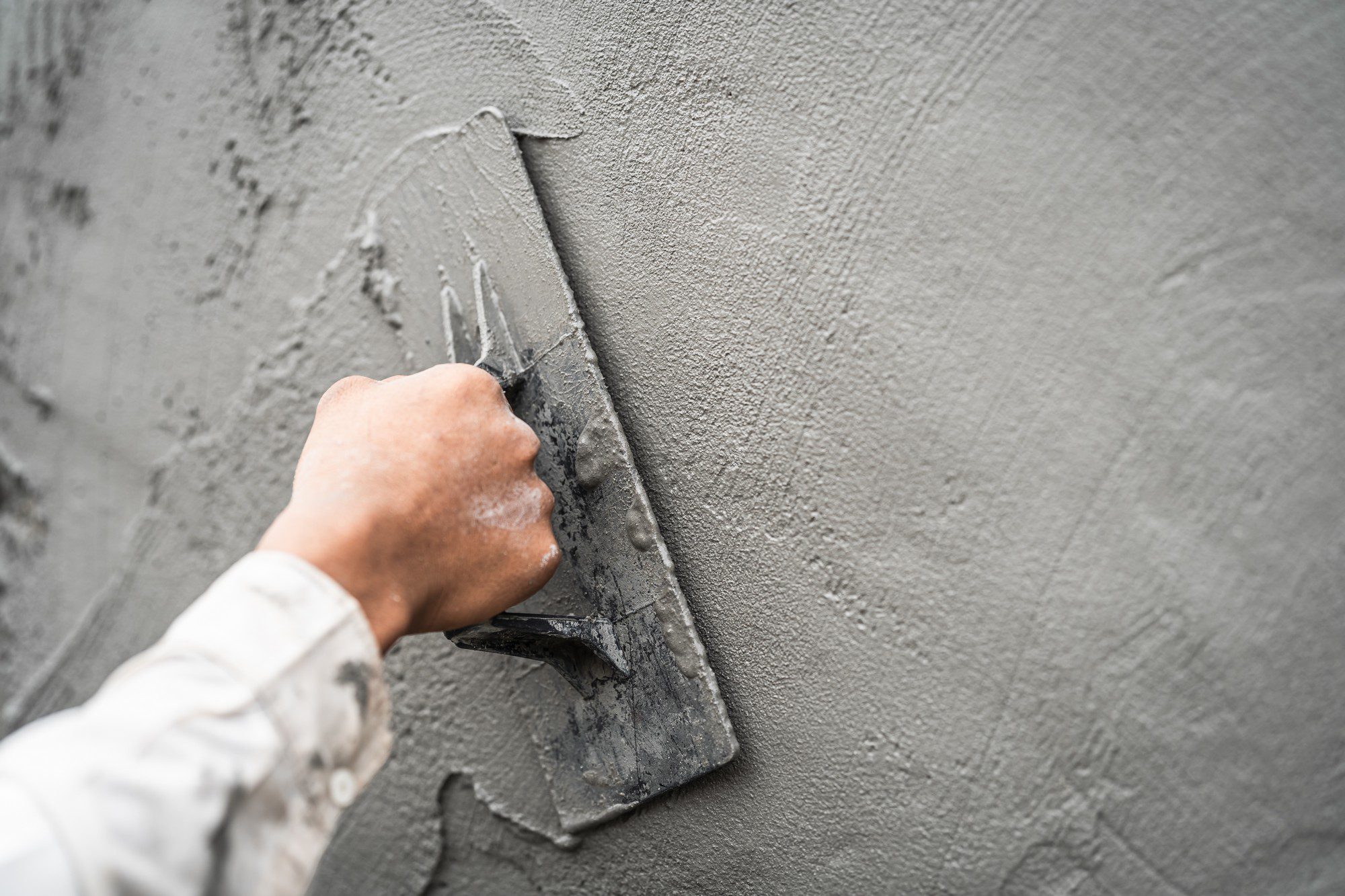 The image shows a person's hand applying a gray construction material to a wall using a trowel. It looks like the process of plastering, a method used in building construction for coating walls and ceilings with a smooth finish before they are painted or wallpapered. The person's hand is covered with some of the material, and they are wearing a long-sleeved shirt that also has splatters of the material on it, indicating that they have been working. The texture of the plaster appears to be smooth and evenly spread, suggesting that the person is skilled in this task.