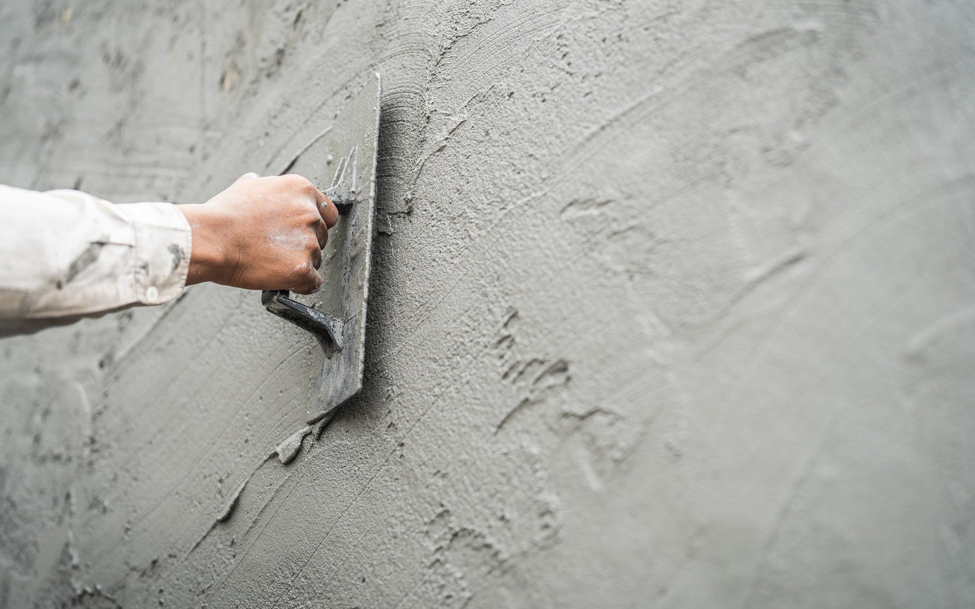 The image shows a close-up view of a person's arm and hand as they use a trowel to apply and smooth out wet cement or plaster on a wall. The process depicted is part of a construction or masonry task, possibly during the finishing phases of wall construction or repair. The person is wearing a light-coloured long-sleeve shirt that has some spots of cement on it, indicating work in progress. The wall appears to be fairly large and uniformly covered with the material, demonstrating part of the plastering process.