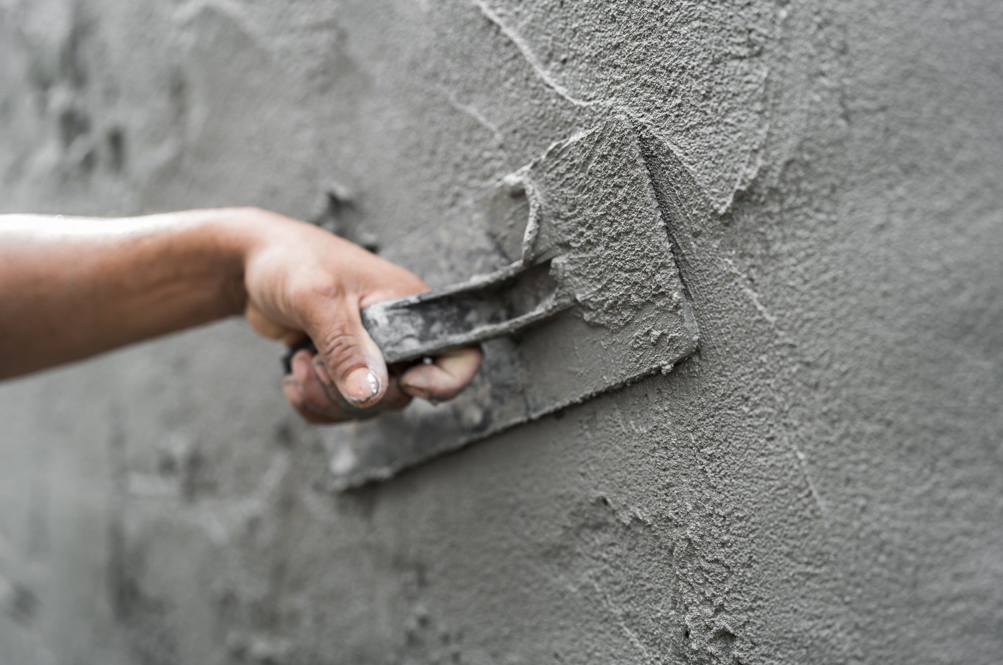 This image shows a person's hand applying a coat of cement or plaster to a wall with a trowel. The trowel is being used to smooth the plaster and the surface is partially covered, indicating the work is in progress. The wall has a rough texture where the fresh plaster has not yet been smoothed out, and some parts have a smoother finish where the trowel has been applied. The person appears to be a construction worker or someone skilled in masonry or plastering.