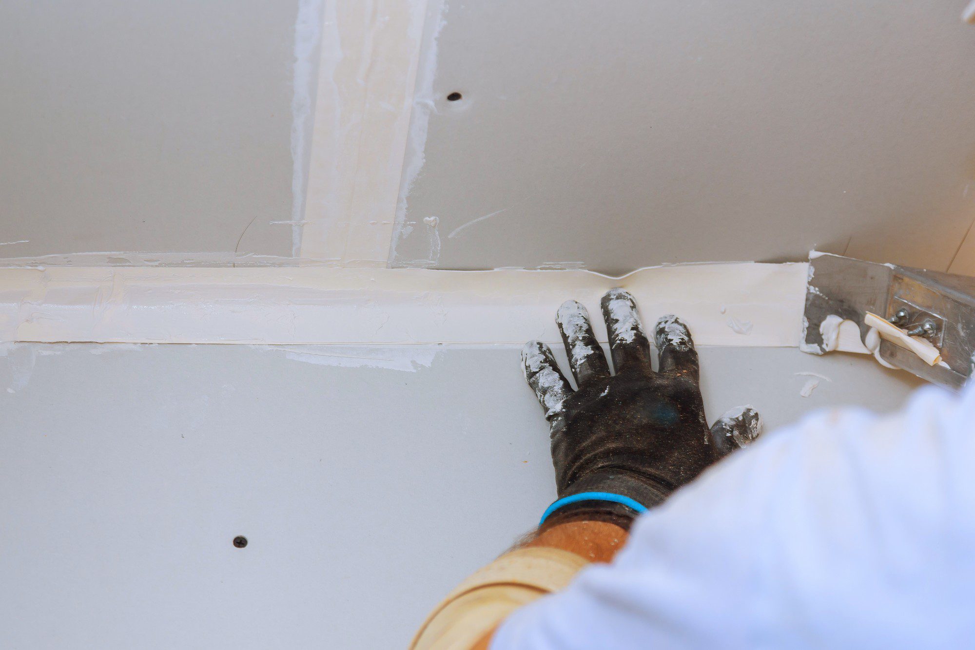 The image shows the corner of a room under construction or renovation. The focus is on a person's hand wearing a black, dirty work glove, which is pressing against a seam where the wall meets the ceiling. It looks like the person is applying or smoothing joint compound, also known as drywall mud, to cover the seam and potentially the head of a drywall screw, which is also visible. A putty knife, which is a tool commonly used for this type of work, is held against the wall just to the right of the hand.

The elements in the picture, such as the unfinished drywall, the joint compound, and the putty knife, suggest that the person is in the process of finishing the drywall. This process includes taping and mudding the seams between drywall panels to create a smooth surface for painting or wallpaper. The presence of excess compound and smudges on the glove and surrounding areas indicates that the work is ongoing.