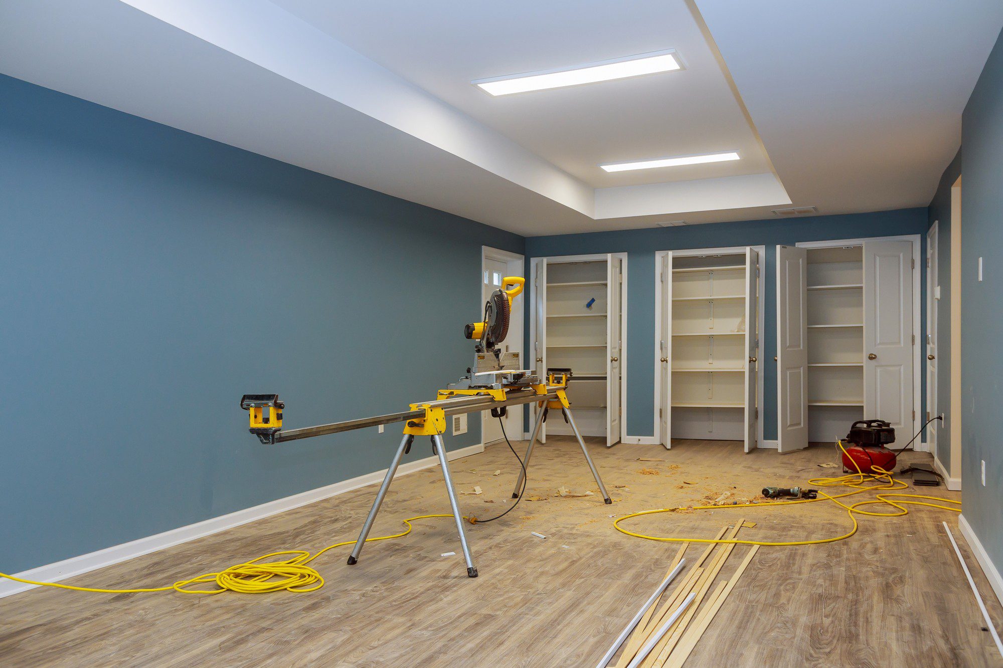 The image shows an interior space undergoing some form of renovation or construction. Notable features in the image include:

1. **Miter Saw**: There is a yellow and black miter saw on a stand with long supports on either side for cutting pieces of wood. This indicates that woodworking is occurring, perhaps as part of installing trim or shelving.

2. **Built-in Cabinets**: On the far side of the room, there's a set of white built-in cabinets with the doors open, appearing to be in the process of construction or installation.

3. **Construction Debris**: Various wood scraps and dust are scattered on the floor, showing active construction work.

4. **Electrical Cords and Tools**: There is a yellow extension cord running across the floor and what looks like a red vacuum cleaner in the background, presumably for cleaning up construction debris. A power drill is also visible on the floor.

5. **Flooring**: The floor is covered with laminated planks that mimic the appearance of wood, suggesting that flooring work might also be part of the renovation.

6. **Lighting**: The room is well-lit with recessed ceiling lights, and there is a feature where the ceiling is raised with additional light fixtures, potentially a tray ceiling.

7. **Colour Scheme**: The walls are painted in a blue-grey tone, and the ceiling is white, which gives a modern and clean appearance.

Overall, the image captures a room in the process of improvement, with various tools