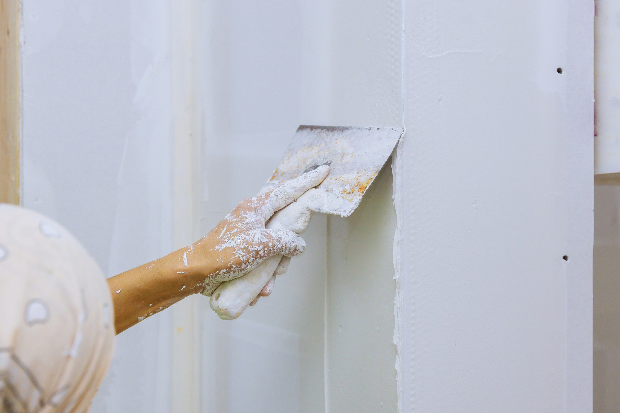 The image shows a close-up of a person's hand holding a plastering trowel against a wall. The wall appears to be made of drywall with visible vertical studs, and it is partially covered with joint compound or plaster. The hand is covered with white dust, likely from the plaster or drywall material, suggesting that the person is in the process of smoothing or applying the joint compound to the wall to create a flat surface for painting or wallpapering. Some of the joint compound is also on the trowel itself. It looks like a typical scene from a wall construction or home renovation project.