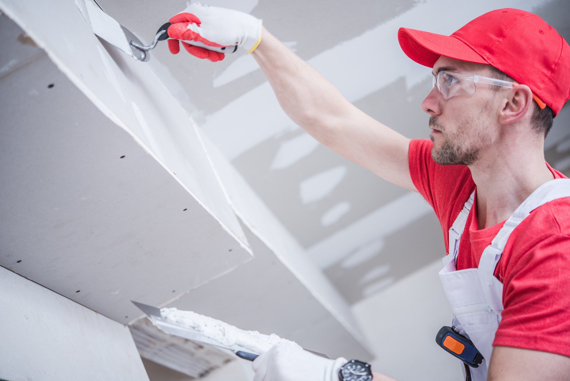 The image shows a person applying joint compound to a seam between sheets of drywall. The individual is wearing safety glasses, a red cap, and a red shirt, complemented by a white apron with pockets, possibly for tools. They are using a drywall taping knife to apply the compound. The person is wearing gloves and appears to be focused on ensuring the joint compound is smooth and even. This is a common step in the process of drywall installation before it can be sanded and painted.