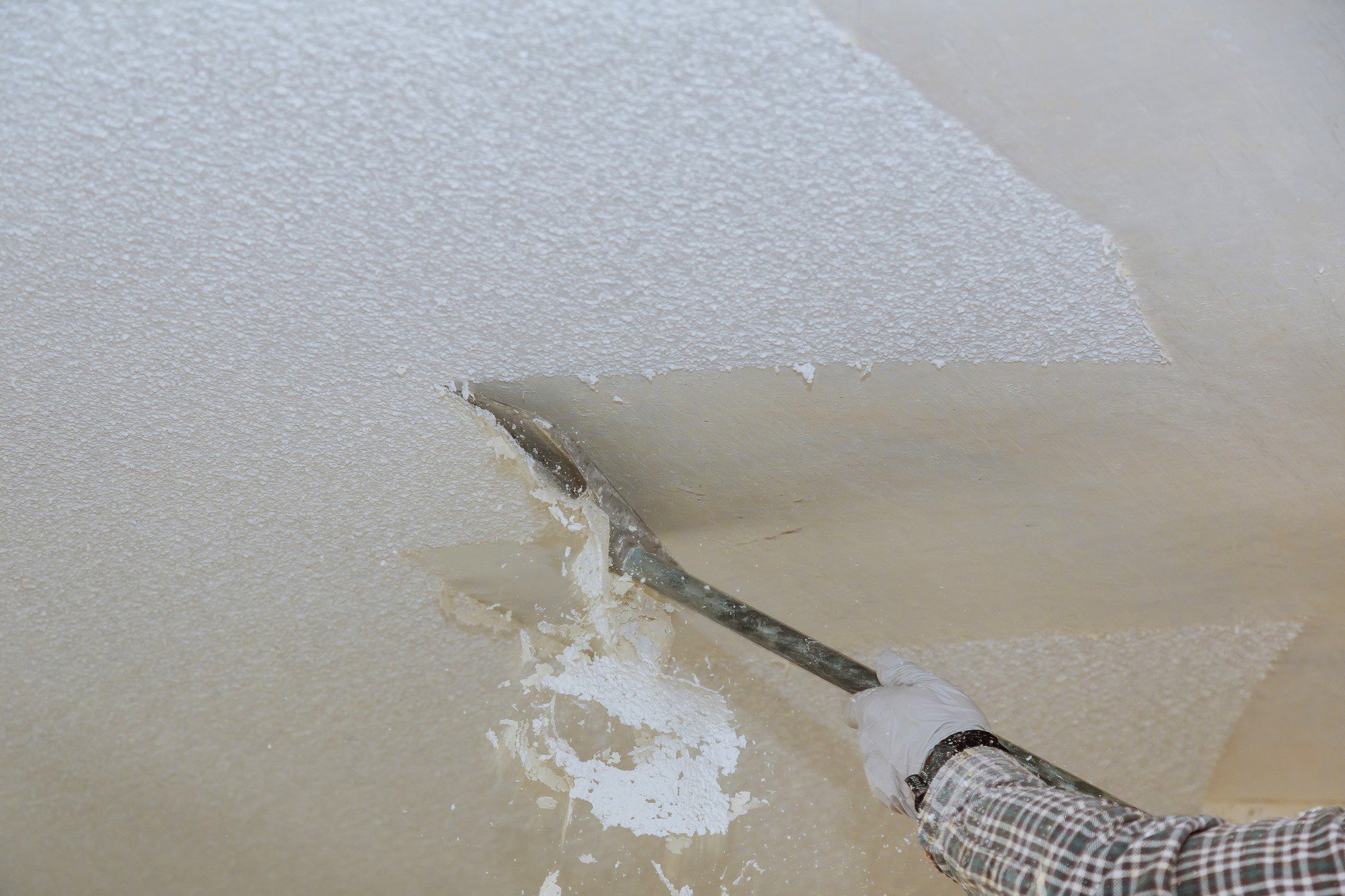 The image shows a person using a floor scraper to remove a layer of material from a concrete floor. The worker is wearing gloves for protection and is in the process of stripping away a white coating, possibly paint, epoxy, or some other finish. You can see where the material has already been removed, revealing the bare concrete surface beneath, and where the coating still remains intact. The tool appears to be a long-handled scraper, which is common in flooring and renovation work to remove flooring materials or finishes.