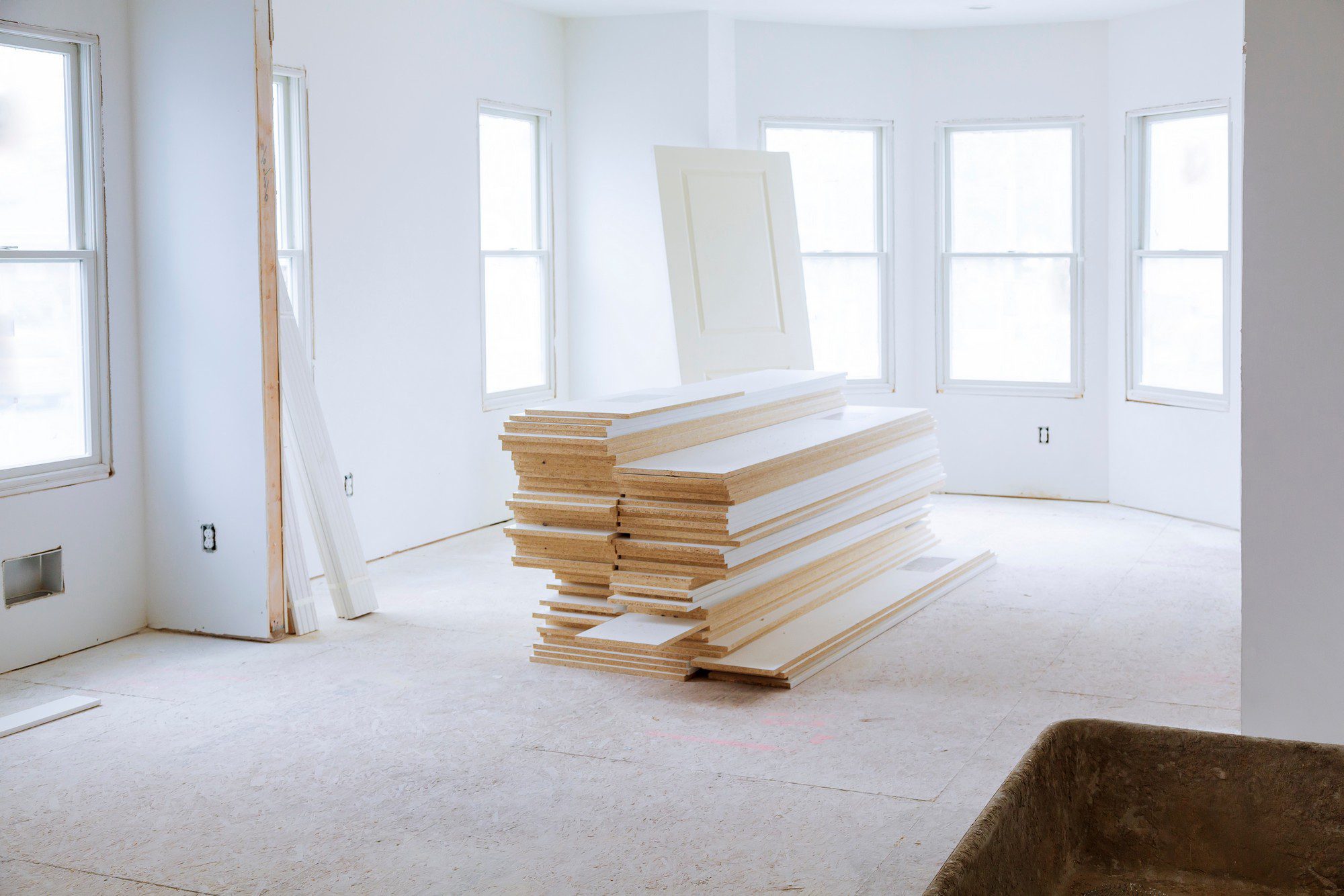 The image shows an interior space under construction or renovation. In the foreground, there's a pile of new, pre-finished door or window trim boards stacked on the floor, awaiting installation. The space has a plywood subfloor and several white walls with multiple windows letting in natural light. Electrical outlets and wall plates are visible, but no cover plates have been installed yet. On the left, there is a lone two-by-four timber leaning against the wall. A construction debris cart or tub is visible in the bottom right corner of the image. The overall appearance suggests that work is progressing but is not yet complete.