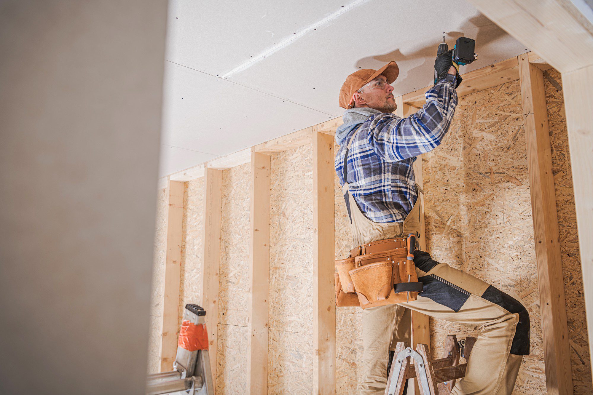 The image shows a person at work on a construction or home improvement project. The individual is wearing a plaid shirt, tan pants, a tool belt, safety glasses, and a baseball cap. They are using a power drill to secure what appears to be a drywall panel to a wooden stud frame. The environment suggests that this is an interior space under construction or renovation, with visible structural timber framing and wood paneling. There is a ladder positioned near the worker, indicating they may have used it to reach the height of the work area. The scene reflects typical construction or carpentry work.