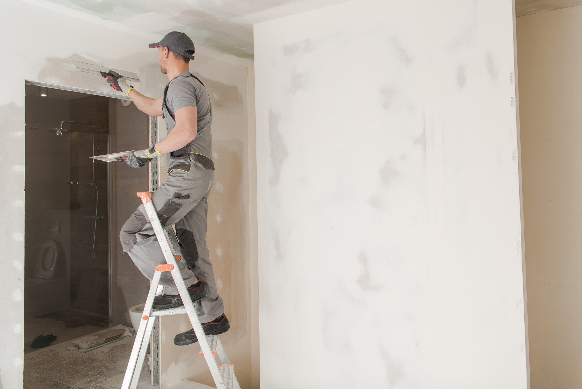 The image shows a person working on a wall inside a room. They are standing on an A-frame ladder and appear to be applying joint compound or plaster to a seam or joint in drywall, as part of the wall finishing process. The person is wearing casual work attire, including a gray shirt, gray pants, and protective gloves, along with a cap. Behind them is what looks like a glass door, possibly leading to a bathroom or shower, given the appearance of tiles seen through the glass. The wall they are working on is not yet finished—patches of compound are visible, suggesting that the wall might soon be sanded smooth and made ready for painting or wallpapering.