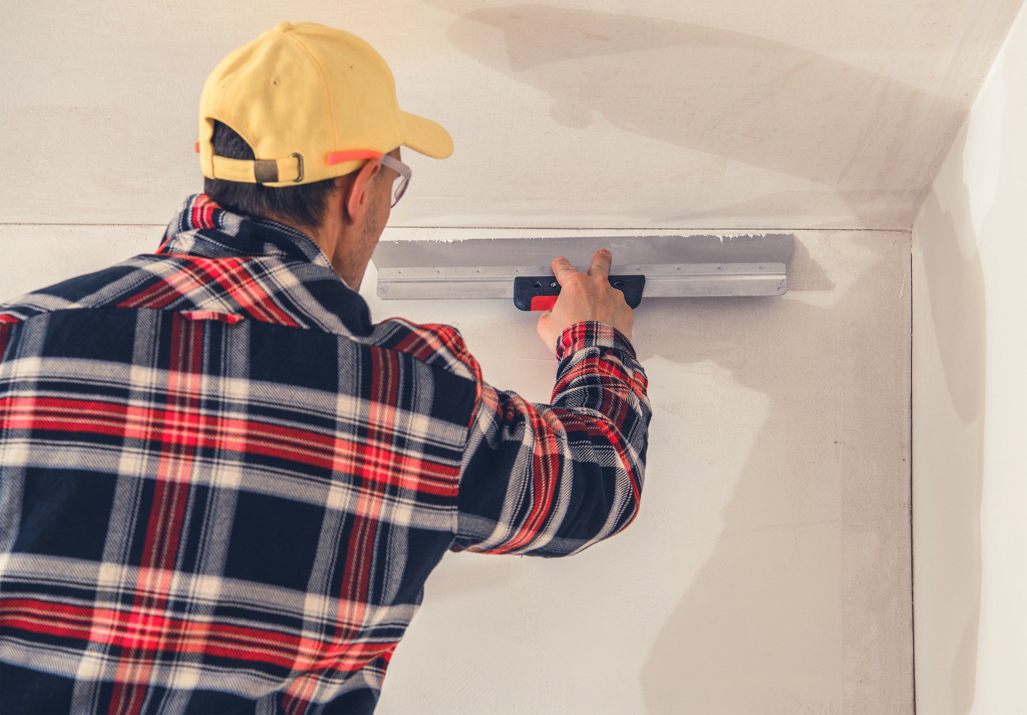 The image shows a person in the process of smoothing a wall surface. The individual is wearing a yellow baseball cap, safety glasses, and a plaid shirt. They are using a long-handled tool, which appears to be a plastering trowel or skim coat trowel, to apply or smooth out plaster or joint compound on a wall. The wall has a rough plaster coat, and it looks like the person might be in the midst of a wall-finishing or construction task. There's noticeable unfinished plaster work around the person, suggesting that the smoothing process is ongoing.