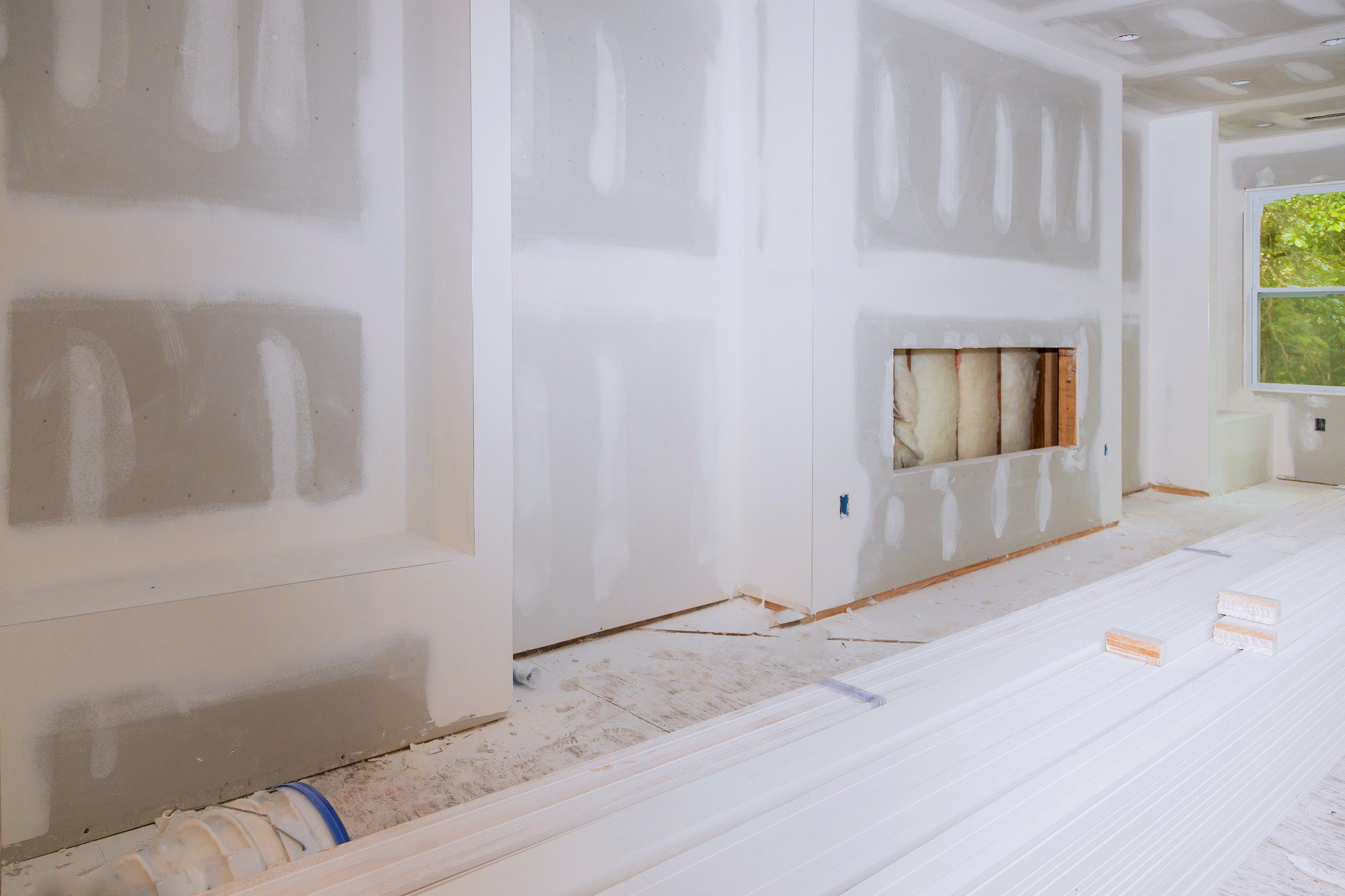 The image shows a room undergoing renovation or construction. The walls are unfinished with visible drywall joint compound applied in vertical and horizontal lines, presumably over the seams between sheets of drywall to create a smooth surface once sanded and painted.

A fireplace has been framed out, but it appears incomplete as it's missing its facade and you can see the insulation behind it. There's a window on the right side letting in natural light, and the ceiling has recessed lighting installed.

On the floor, there are several lengths of white primed baseboard trim and two blocks of wood, which will likely be used in the finishing of the room. The floor is covered in construction debris and dust. The blue tape on the floor suggests that some finished surfaces are being protected during the construction process. Overall, the picture depicts a work in progress, with various stages of construction evident.