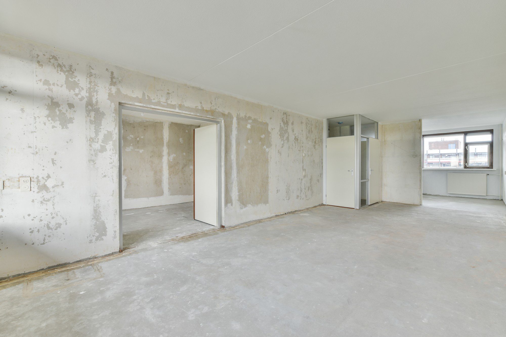 The image depicts an unfurnished and unrenovated interior room of a building or flat. The walls are in a state of disrepair with peeling paint and some exposed areas where the underlying concrete or plaster is visible. There is a built-in closet with its door open, and an additional open doorway leading into another space. The floor appears to be concrete and is bare with no carpeting or other flooring material installed. There's a radiator under the window, suggesting that the building has central heating, and an electrical outlet on the left wall. The ceiling is white and looks to be in better condition than the walls. The window provides a view outside where other buildings can be seen, indicating an urban setting. Overall, the room looks like it requires renovation before being habitable.