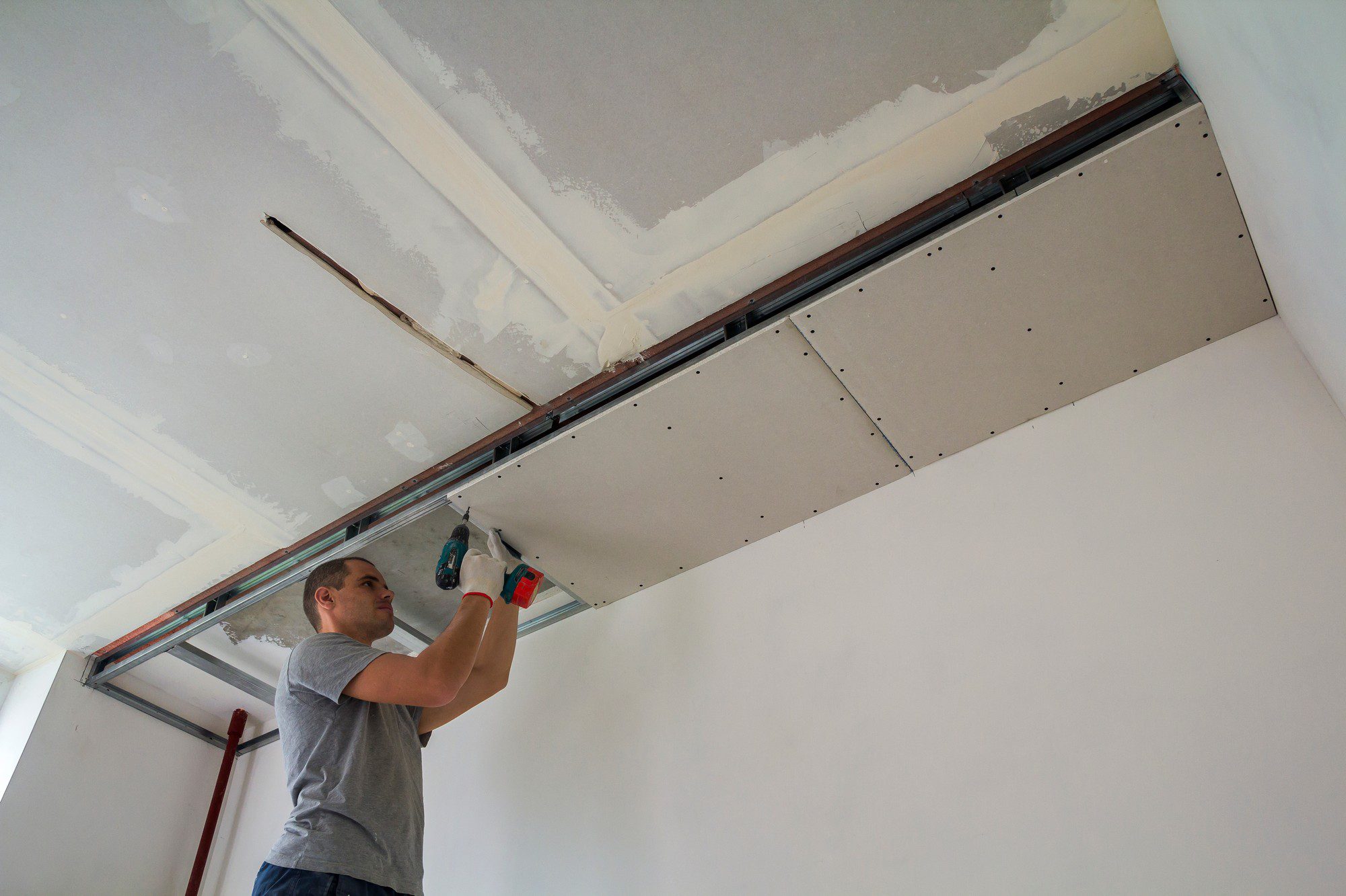 The image shows a person installing drywall or plasterboard on a ceiling. He is using a power drill or screw gun to secure the drywall to the metal framing that has been put in place to support the ceiling structure. The ceiling appears to be under renovation or construction, as indicated by the unfinished surfaces and the presence of tools and materials. The drywall sheets are partially installed, with some edges still untaped, indicating that the work is in progress. The person is wearing casual clothing and seems focused on the task at hand.