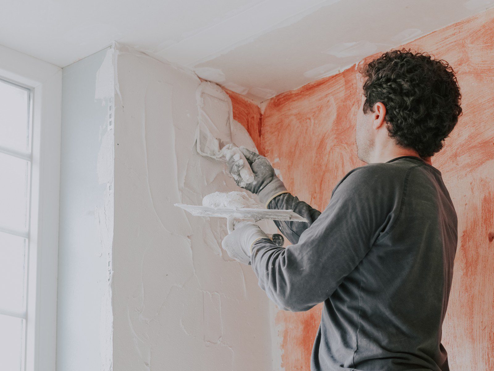 The image shows a person applying joint compound, often referred to as "mud," to a wall with a putty knife. The person is likely in the process of drywall finishing or plastering to prepare the wall for either painting or wallpapering. They are wearing gloves for protection and seem to be applying a smooth layer of the material to fill in gaps and create a smooth surface on the wall. The red-brown colour on the wall indicates an underlayer of material, which could be a different type of wall treatment, potentially something like a bonding agent or primer, that was applied before the joint compound. The environment looks like a home renovation or construction setting.