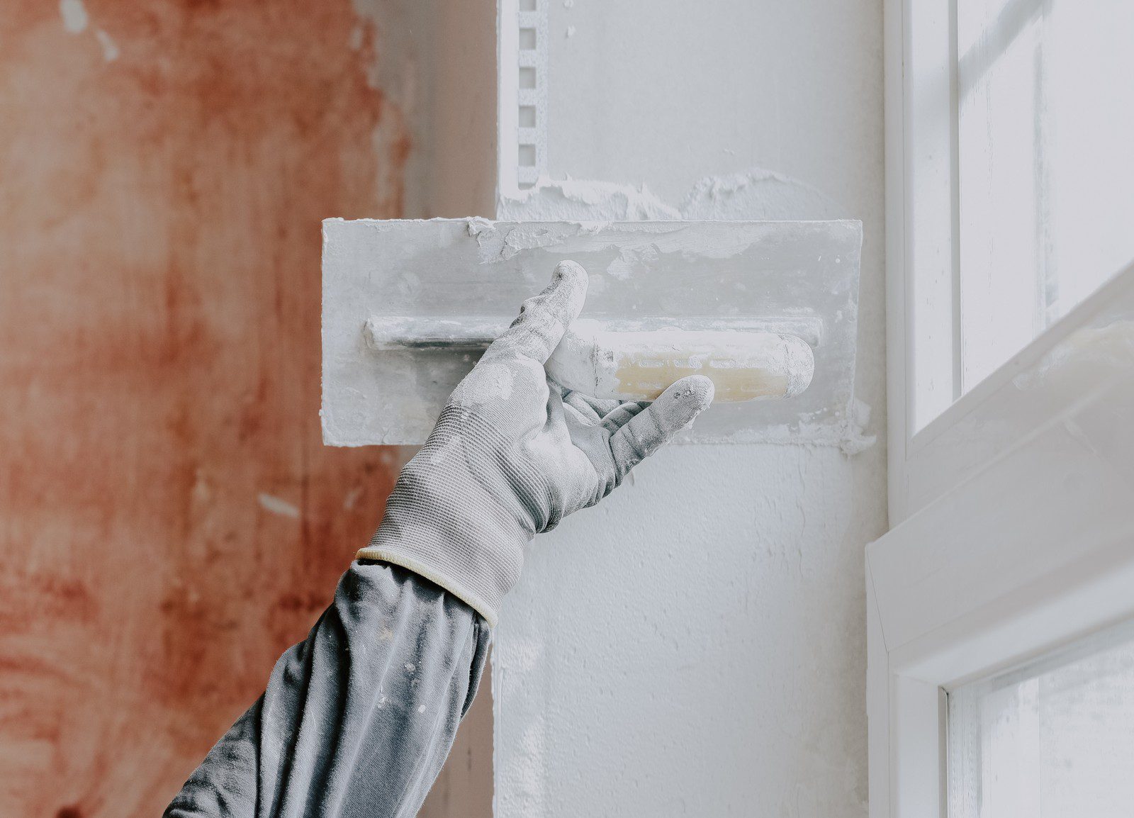 The image shows a close-up of a person's hand applying plaster to a wall with a plastering trowel. The individual is wearing a grey work glove and is in the process of smoothing the plaster or joint compound over the surface near a window, as part of a wall repair or finishing job. You can see the white plaster on the trowel and some has been spread on the wall. The surrounding area suggests it might be a construction or renovation site.