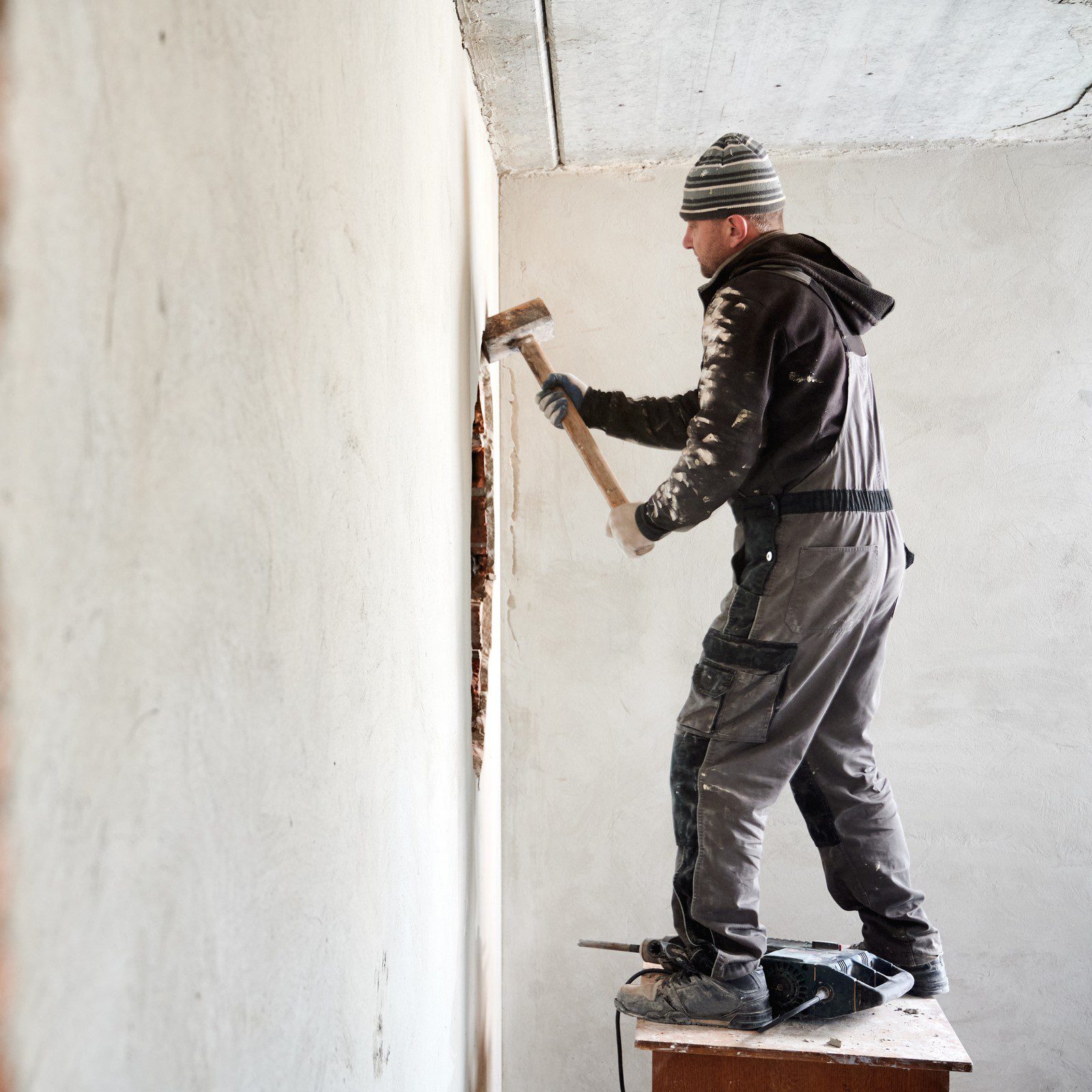 The image shows a person engaged in some form of manual labour, likely construction or renovation work inside a building. The person is standing on a makeshift platform made of a wooden board resting on what appears to be a small table or bench, which is itself on top of another flat surface, probably to reach a higher part of the wall.

This individual is wearing work clothing, including a dark jacket, gray overalls, gloves, and safety shoes. On their head, they have a striped beanie. They are wielding a hammer in their right hand and are in the process of using it on a wall that has a partially exposed area, indicating that old materials are being removed, or an opening is being enlarged or shaped.

The room they are in has an unfinished look, with bare walls that have not been fully smoothed or painted, and the lighting suggests that there is natural light coming from outside the frame, possibly from a window to the right. There is a sense of refurbishment or remodeling in progress.