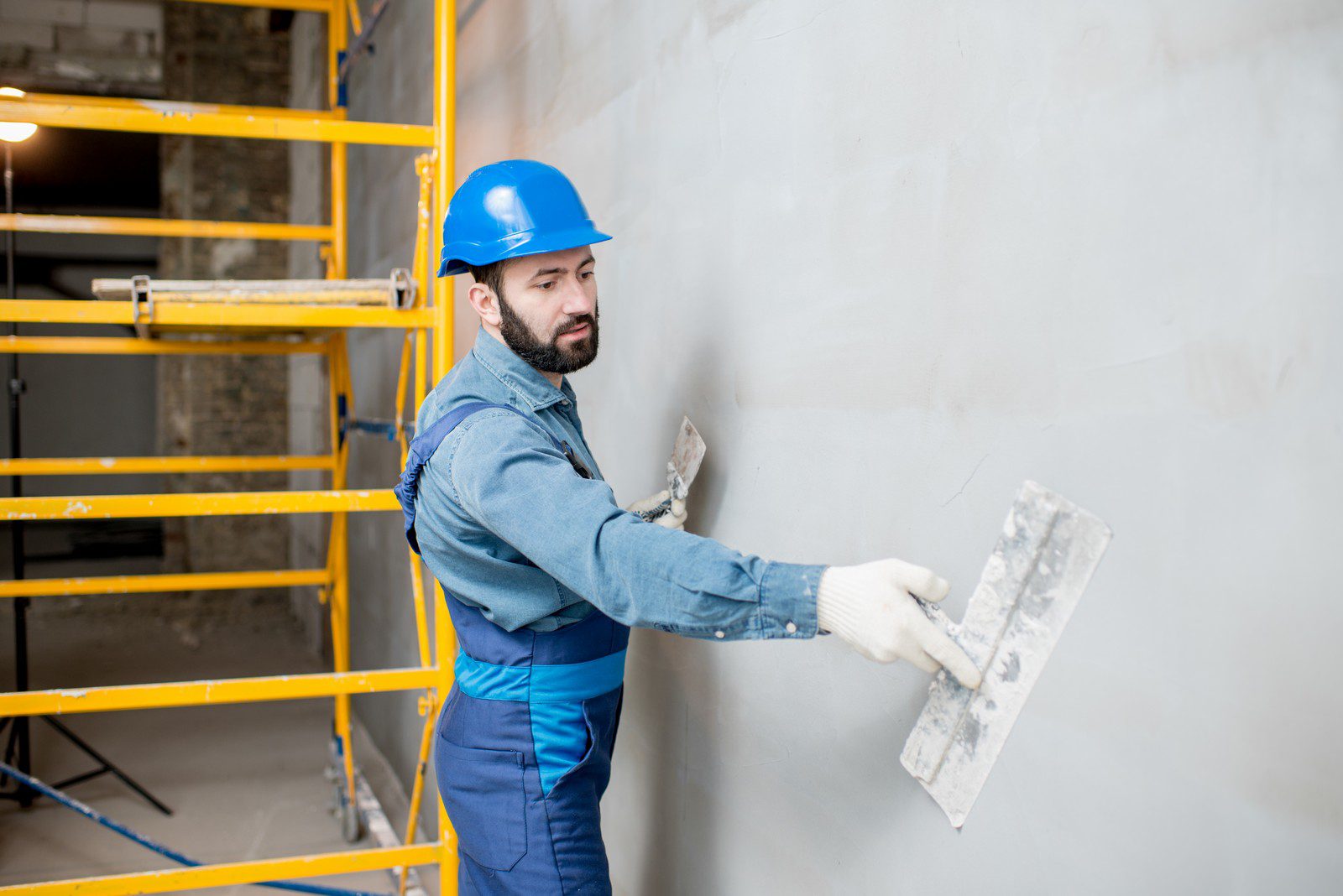 The image shows a man dressed in work attire including a blue hard hat, a denim shirt, and blue overalls. He appears to be a construction worker or plasterer, engaged in a task where he is applying or smoothing out material on a wall with a trowel. In the background, there's a yellow scaffolding structure, indicating that this is a construction or renovation site. The wall the man is working on is gray, likely from the material he is applying or smoothing. His expression seems focused on the task at hand.