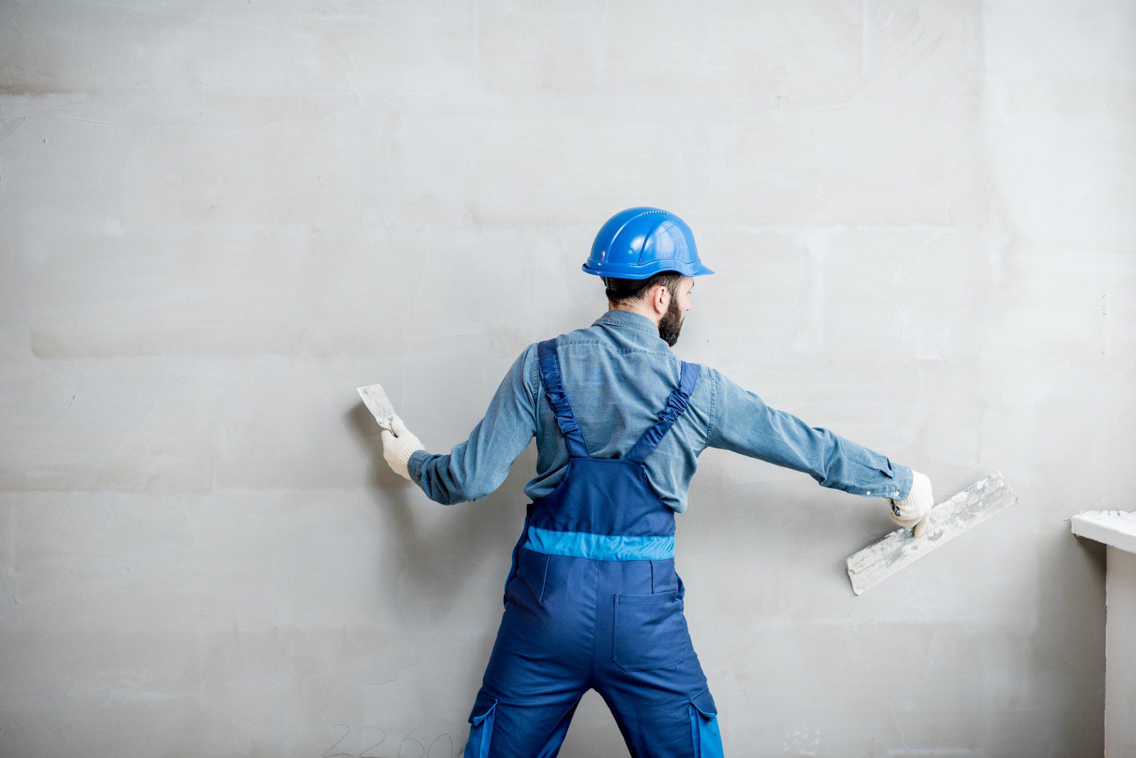 In the image, you see a person wearing a blue hard hat and a blue work uniform with braces, involved in a construction or plastering task. The person is using what looks like a plastering trowel in each hand to apply or smooth plaster on a wall. The wall is partially plastered, indicating that the work is in progress. The individual seems to be in a working pose and focused on the task at hand. There appears to be a slight shadow of the person on the wall, indicating that the lighting is coming from the front. The background suggests an indoor work environment, probably a room that is being renovated or constructed.