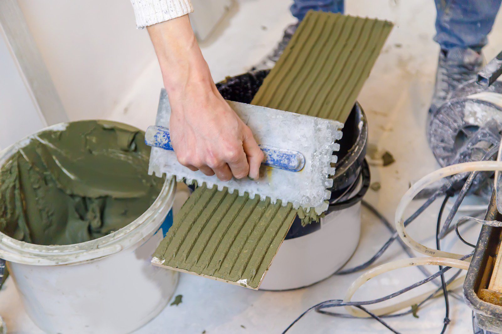 The image shows a construction or renovation setting, where a person's hand is holding a notched trowel with a greenish adhesive material on it, usually used for laying tiles. The material on the trowel has been combed to create ridges, which is a standard technique for ensuring a strong bond when setting tiles. A bucket partially filled with the same adhesive material is also visible, as is an electric drill with a mixing paddle attachment, likely used to mix the adhesive to its proper consistency. The person appears to be in the process of working with these materials, probably preparing to install tiles.