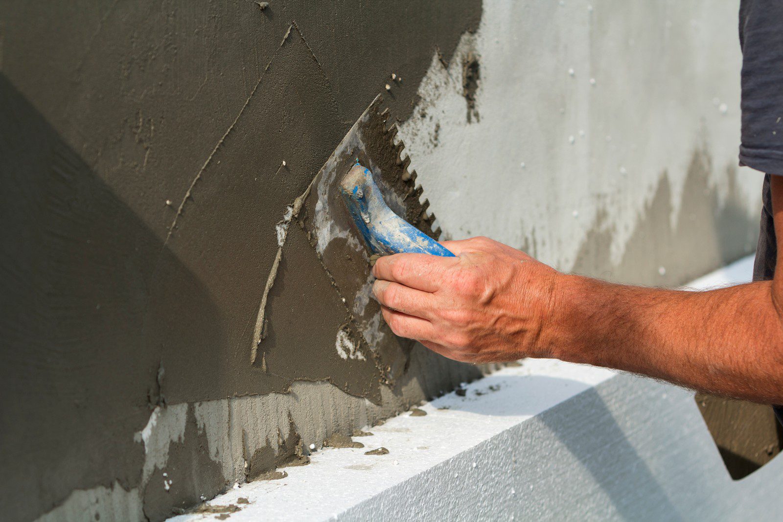 The image shows a person applying plaster or stucco to a vertical wall surface. The individual is using a trowel to spread and smooth the mixture onto the wall. The trowel appears to have notches, which are commonly used to create a textured pattern in the plaster or to ensure an even layer of adhesive when laying tiles. The wall looks partially covered with the fresh material, and some areas are still bare, showing the different stages of the plastering process. The person's hands are clearly visible as they handle the trowel, and it looks like a close-up action shot of the plastering work.