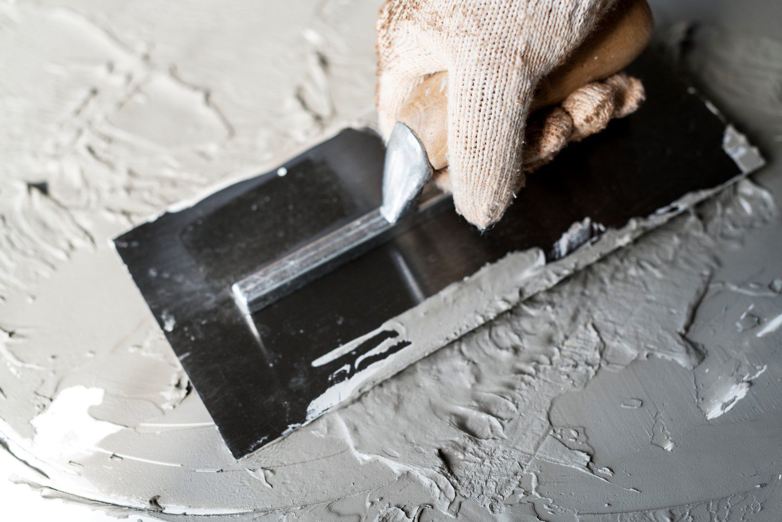 The image shows a close-up of a person's hand wearing a fabric glove while using a metal putty knife or spatula to apply a gray compound, which appears to be some kind of putty, plaster, or joint compound, onto a flat surface. The flat surface might be part of a wall, ceiling, or board that is being prepared or repaired, as this type of material is often used for filling cracks, smoothing surfaces, or creating finishes in construction and renovation projects. The scene suggests a focus on craftsmanship, attention to detail, and the physical aspect of manual work.