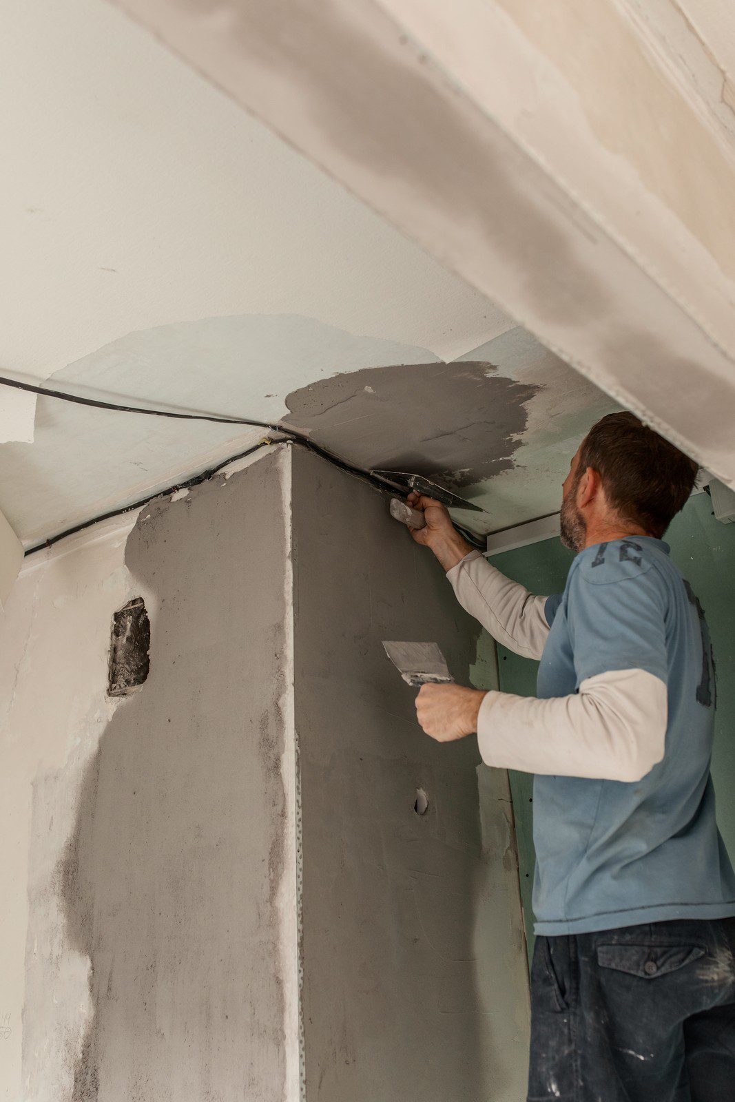 This image features a person who appears to be a tradesman applying a plaster or joint compound to a wall or ceiling with a plastering trowel. The work involves smoothing the surface for a finished look, possibly as part of a construction, renovation, or repair project. The person is in the process of ensuring the material is applied evenly and is likely skilled in plastering or drywall techniques. The plaster or compound is gray, and there is a noticeable difference in colour between the applied area and the rest of the white ceiling or wall, indicating the work in progress. There's also some exposed wiring, hinting that other construction or electrical work might also be occurring in this space.