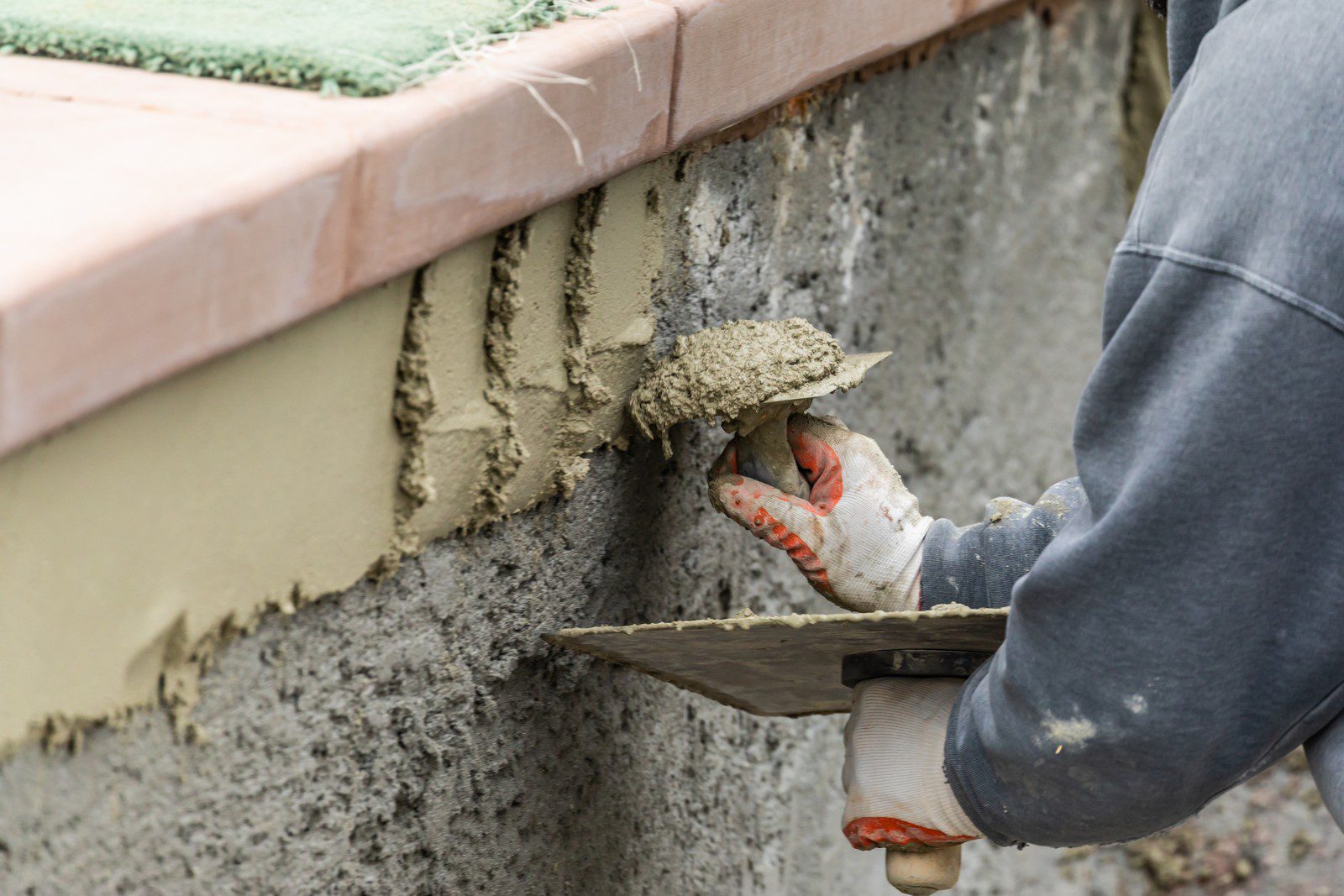 The image shows a person applying wet cement or mortar to a wall with a trowel, a common tool used in masonry. The individual is wearing a long-sleeve shirt and protective gloves, indicating that they are adhering to safety practices while working with construction materials. The person is in the process of smoothing or spreading the mortar onto the wall surface, which appears to be part of a construction or repair process. The wall is partially covered with a new layer of mortar, and you can see the rough surface of the wall below where the new layer has not yet been applied.