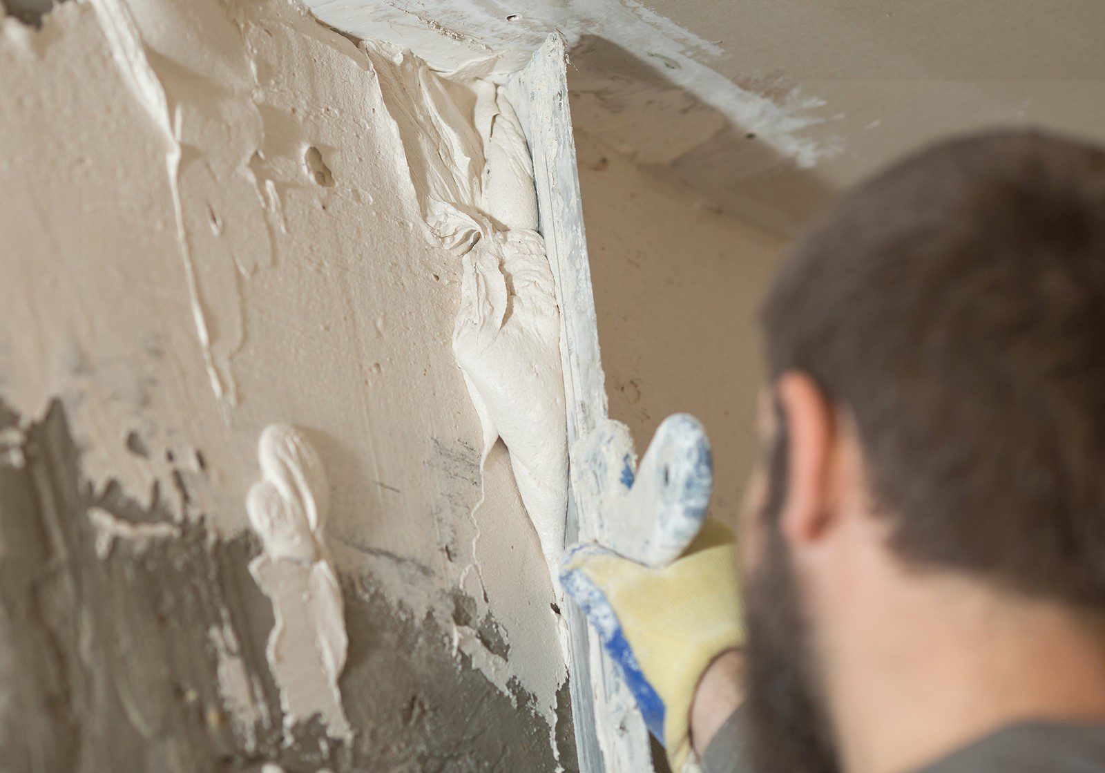 The image shows a person in the process of applying plaster or spackling paste to a wall or corner, likely repairing it or preparing it for painting. The individual is wearing a glove and holding a putty knife, commonly used for spreading and smoothing plaster. The area has patches of uneven, wet plaster that have not yet been smoothed out. The focus is on the work and not on the individual's face, as the person's back is to the camera and their head is turned away.