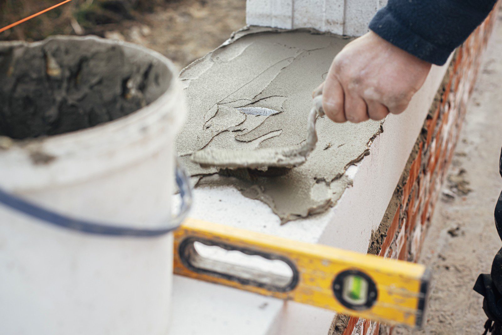 The image depicts a construction worker engaged in the installation of white masonry blocks, which are typical of modern building projects. The worker is seen in a close-up shot, focused intently on the task at hand. He wears essential safety gear, including a yellow hard hat, high-visibility vest, protective gloves, and safety glasses. The worker is in the process of applying mortar with a trowel, ensuring that the blocks are level and securely placed. The white masonry blocks are neatly aligned in a partially-built wall, showcasing a clean and precise construction technique. The background includes additional building materials and construction tools, indicating an active building site. The overall scene is characterized by a sense of meticulous craftsmanship and adherence to safety standards. The lighting is natural, suggesting that the work is taking place during daylight hours.